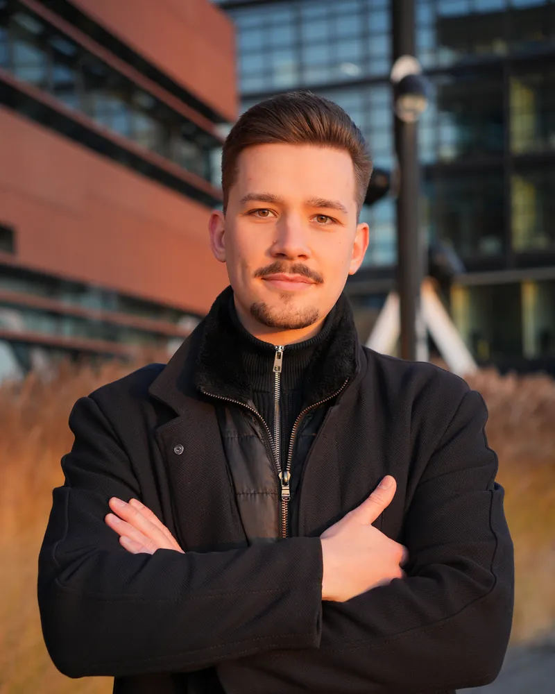 Tom Falz im dunklen Mantel mit verschränkten Armen vor einem Bürogebäude in der Abendsonne